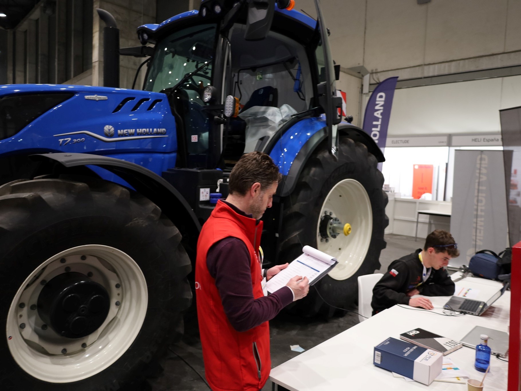 A large blue New Holland T7.300 tractor is displayed indoors at an exhibition or trade show. In the foreground, a man wearing a red vest is holding and writing on a clipboard, while another person is seated at a white table working on a laptop. Various banners and booths are visible in the background.
