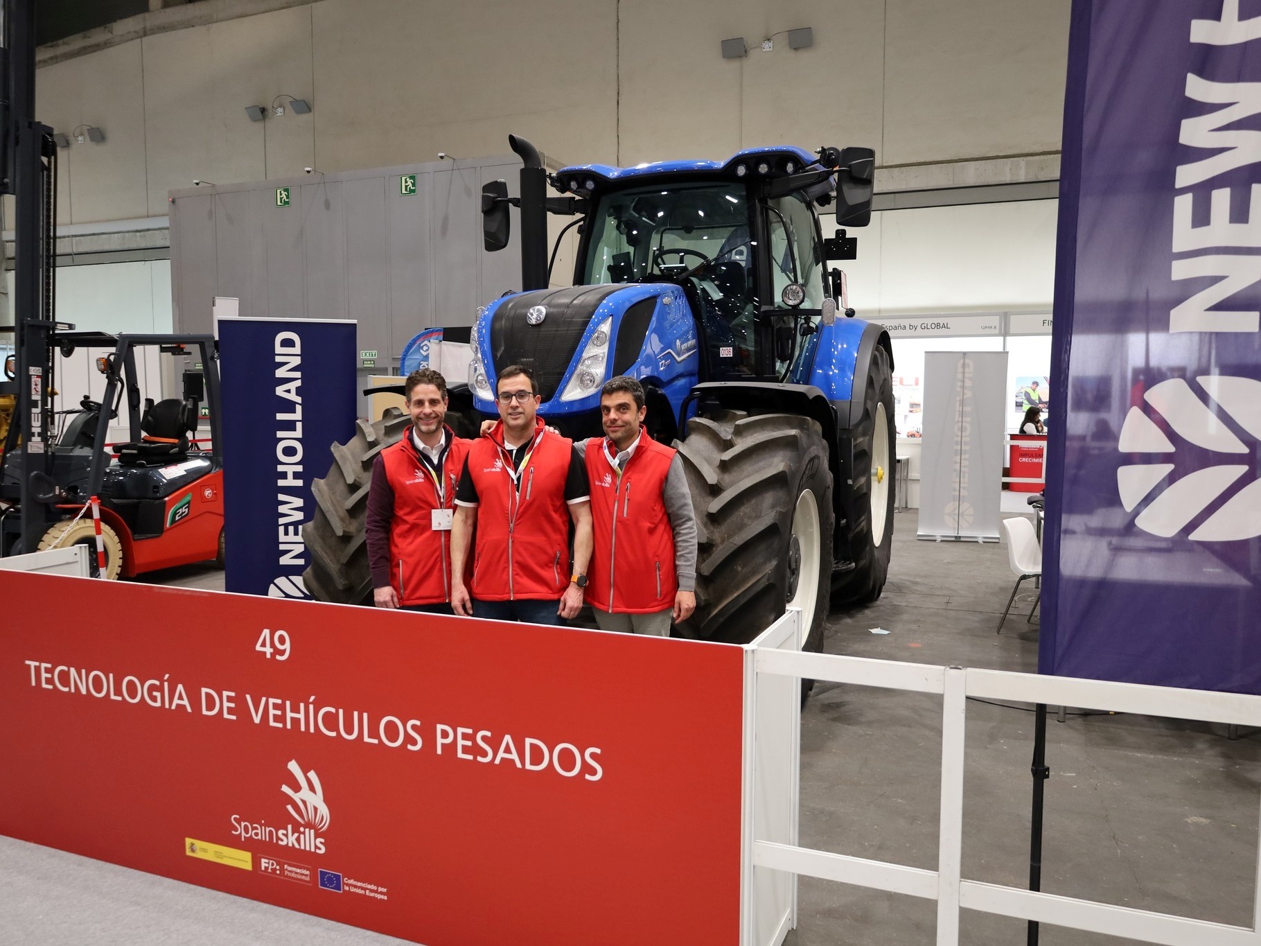 Three men wearing red vests standing in front of a large blue New Holland tractor at an indoor exhibition booth labeled "Tecnología de Vehículos Pesados" with SpainSkills branding.