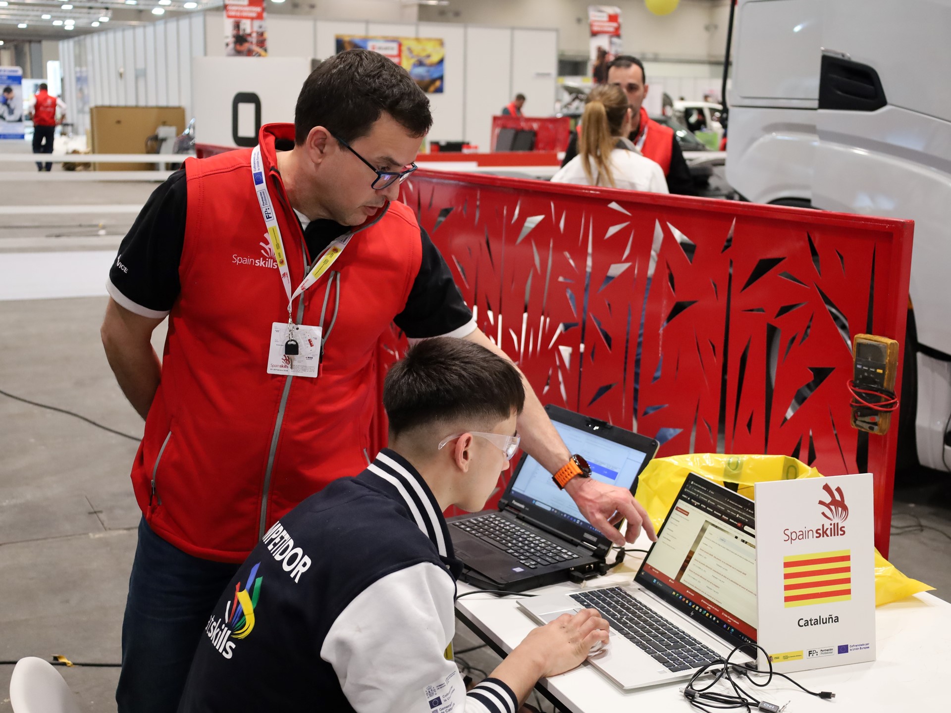 Two men working together on laptops at a SpainSkills event in an indoor workshop setting, with one man standing and pointing at the laptop screen while the other sits and types. A sign on the table displays the SpainSkills logo and the word "Cataluña" with the Catalonia flag. The background shows other participants and equipment in a spacious, well-lit area.