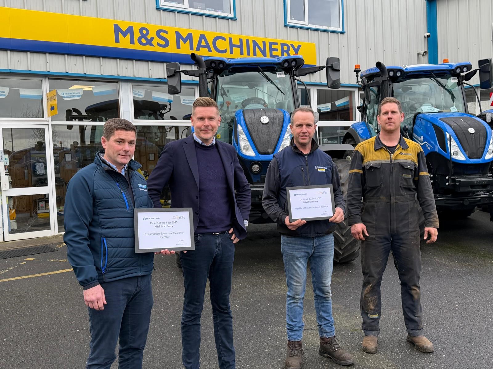 Four men standing in front of two blue tractors outside a building with a yellow and blue "M&S Machinery" sign. Two of the men are holding framed certificates, and all are dressed in casual or work attire. The scene appears to be an award presentation or recognition event.