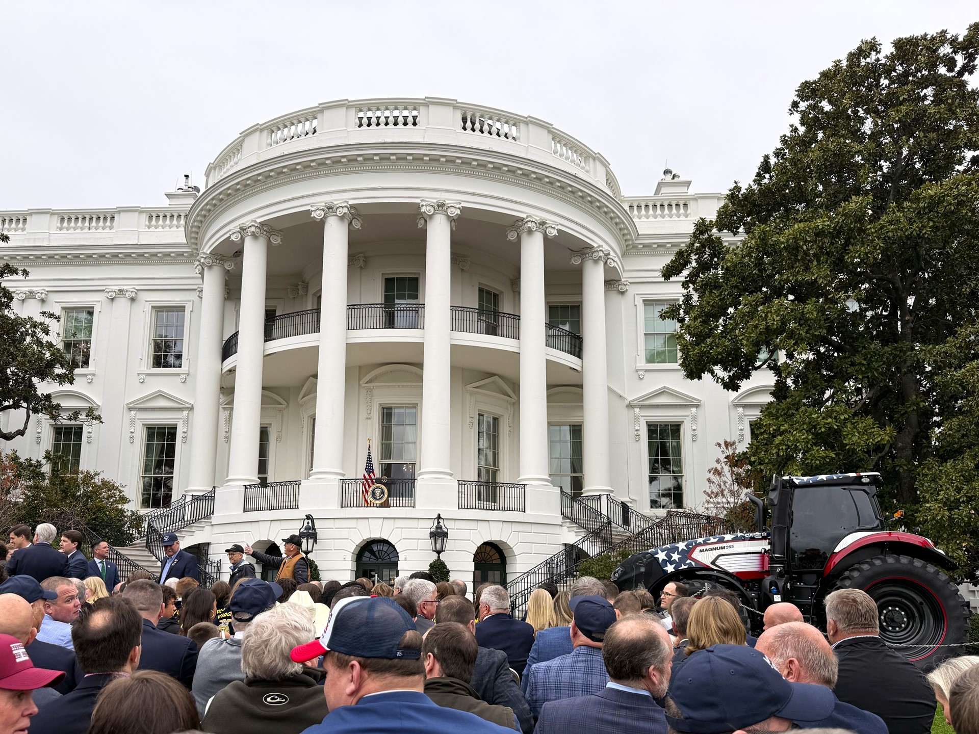A large crowd of people gathered in front of the South Portico of the White House, with a tractor decorated in red, white, and blue parked to the right side near a large tree, under an overcast sky.