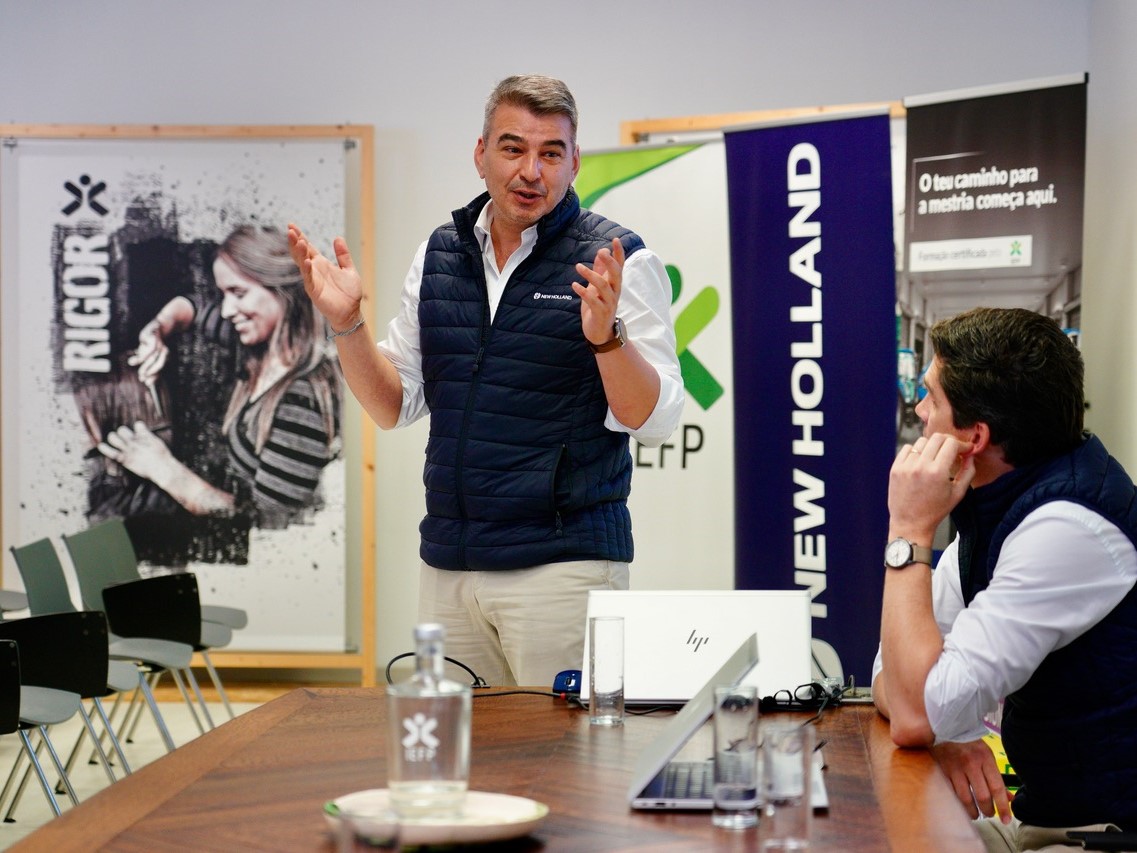 Two men in a meeting room, one standing and speaking with expressive hand gestures while the other sits at a table listening attentively. Both are wearing navy blue vests over white shirts. The table has laptops, glasses of water, and a water bottle. In the background, there are banners with the logos "NEW HOLLAND" and "RIGOR," and a poster featuring a woman working with her hands. The setting appears to be a professional or corporate environment.