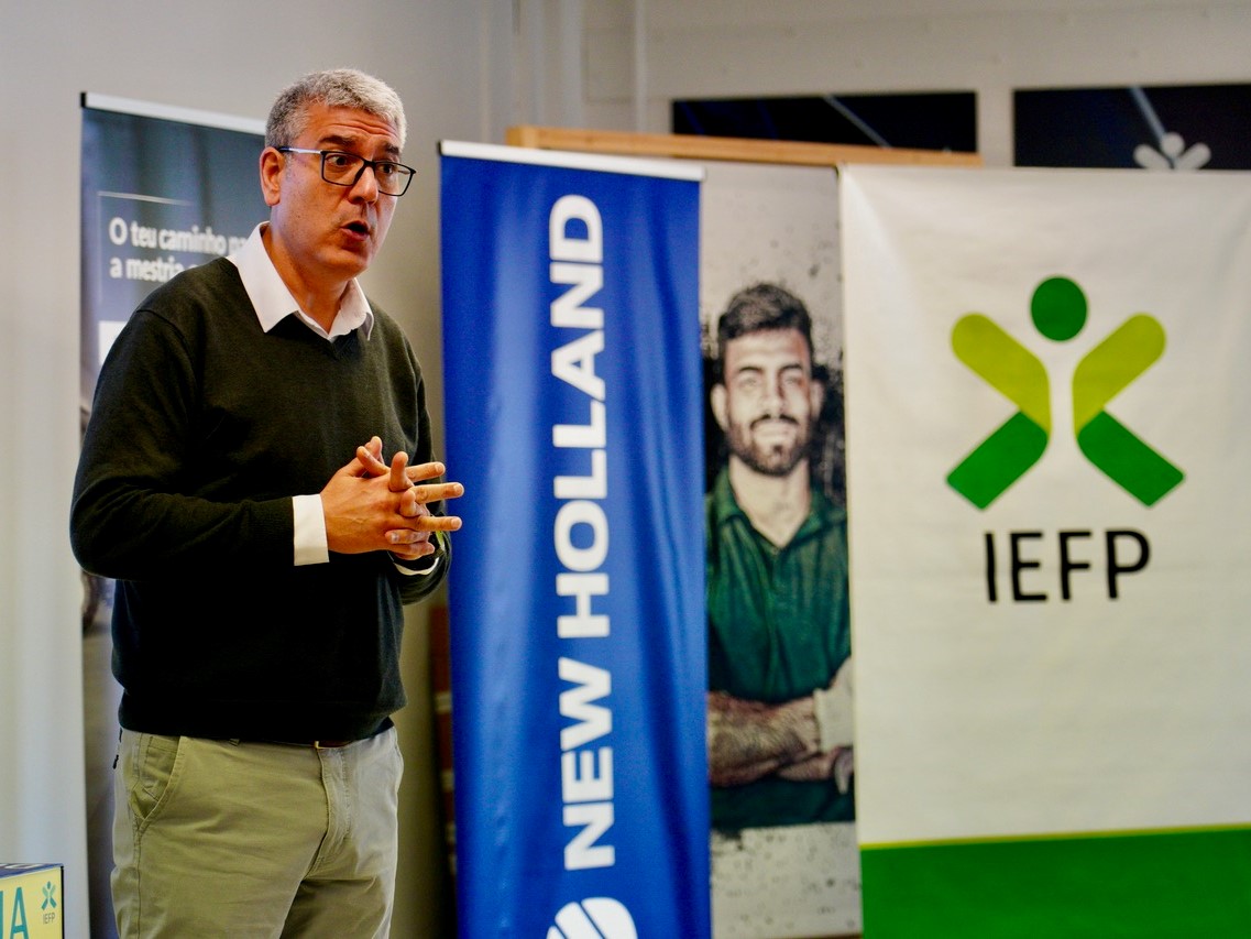 A man with gray hair and glasses, wearing a black sweater over a white collared shirt and beige pants, is speaking or presenting indoors. Behind him are three vertical banners, one blue with the text "NEW HOLLAND," another with a photo of a man with crossed arms, and a white banner with a green and yellow logo and the text "IEFP.