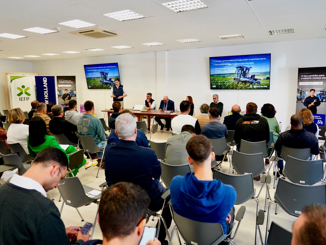 A group of people attending a presentation or seminar in a conference room. A speaker stands at the front near two large screens displaying slides with agricultural machinery images and text. Several panelists are seated at a table beside the speaker. Attendees are seated in rows facing the front, some taking notes or using their phones. Banners for IEFP and New Holland are visible in the background. The setting appears professional and focused on agricultural or farming topics.