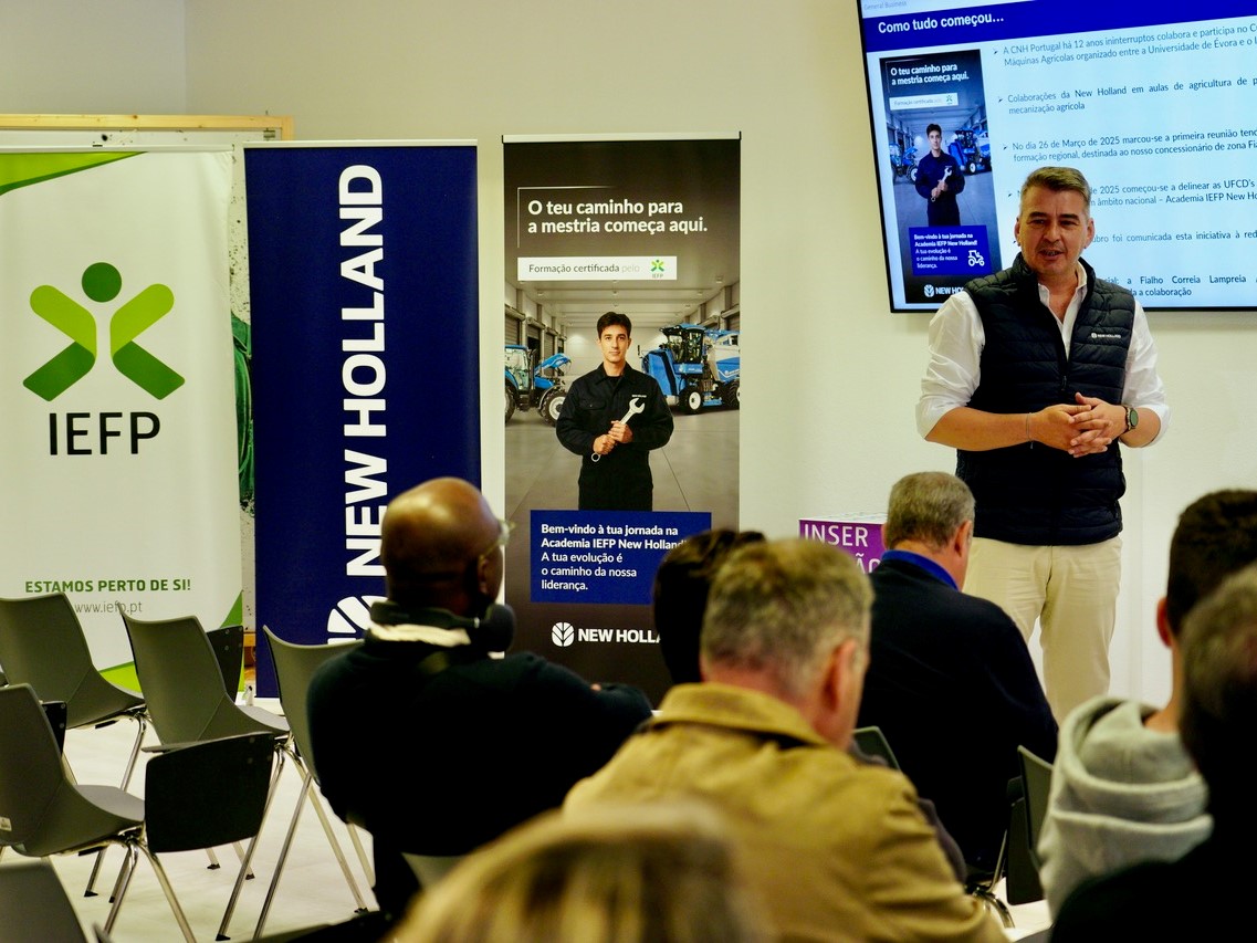 A man in a white shirt and black vest is giving a presentation to an audience seated in a room. Behind him are banners for IEFP and New Holland, and a screen displaying information in Portuguese about a training course and collaboration between IEFP and New Holland. The audience is attentively listening, and the setting appears to be a professional or educational event focused on agricultural machinery training.