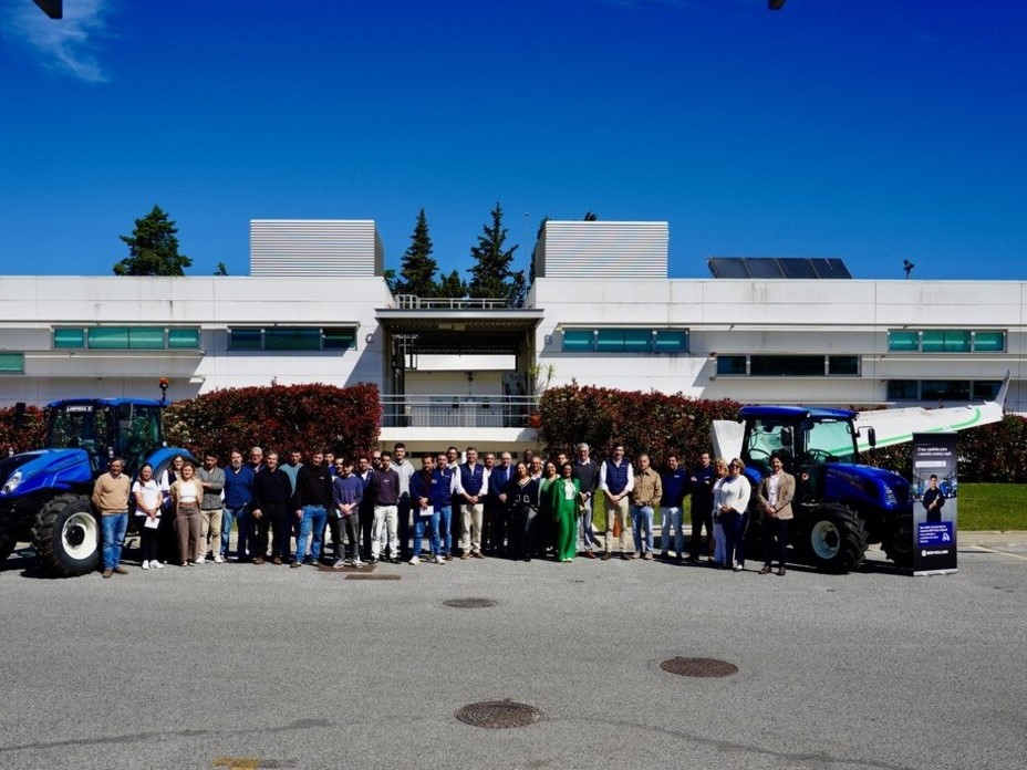 A large group of people standing in front of a modern white building with green bushes and trees in the background. Two blue New Holland tractors are positioned on either side of the group, with New Holland banners next to each tractor. The sky is clear and blue, indicating a sunny day.