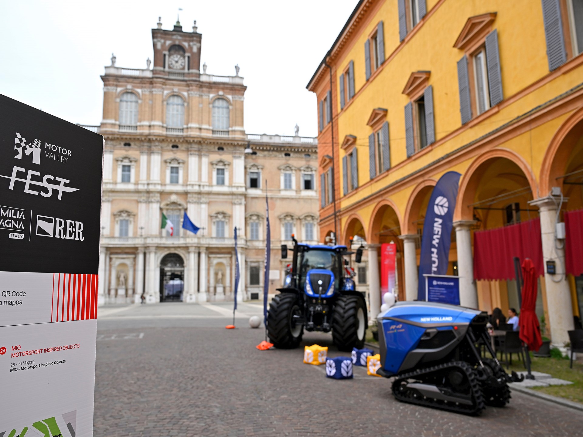 Outdoor exhibition of New Holland agricultural machinery including a blue tractor and a tracked vehicle displayed on a cobblestone street in front of historic buildings with arches and a clock tower in the background, during the Motor Valley Fest in Emilia Romagna, Italy.