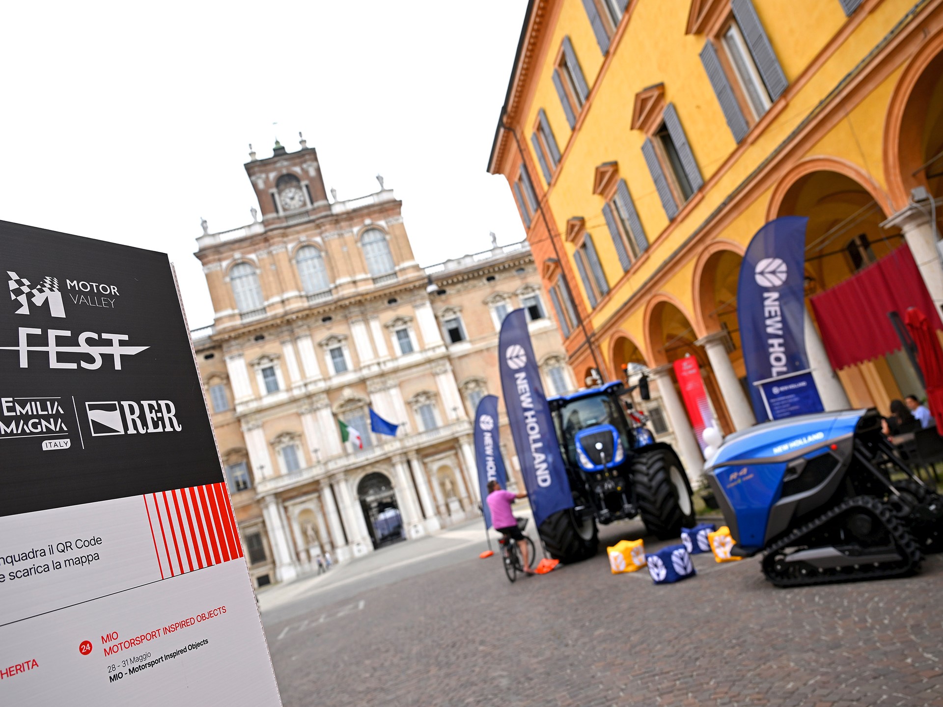 Outdoor event setup in a historic Italian square featuring a Motor Valley Fest sign with logos of Visit Emilia Romagna and RER, alongside New Holland branded agricultural machinery including a blue tractor and a blue tracked vehicle, with a person on a bicycle nearby and classical architecture in the background.