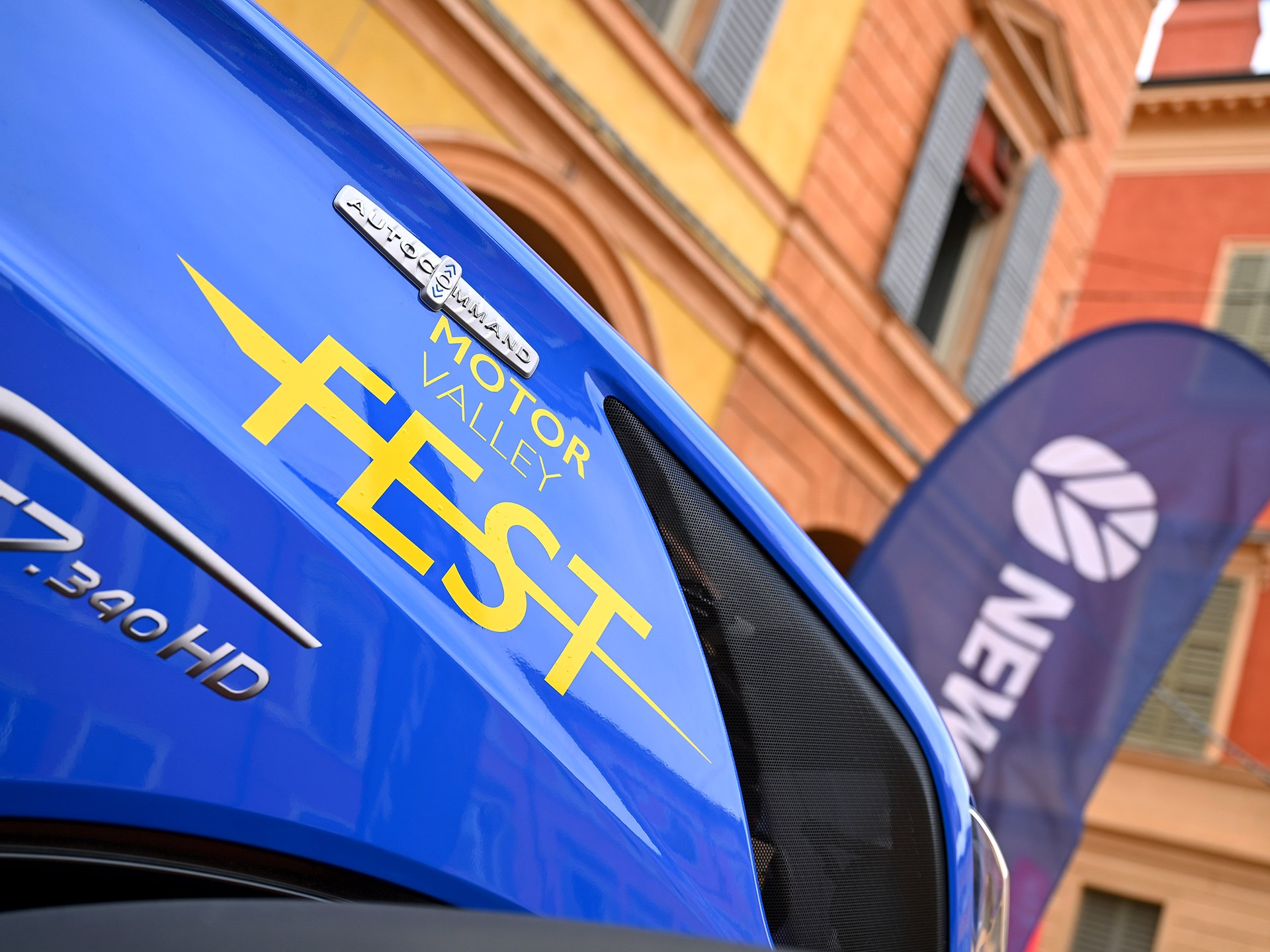 Close-up of a blue vehicle with yellow text "Motor Valley Fest" and model designation "T7.340 HD" on its side, set against a background of colorful buildings and a blurred flag with the word "New" visible.