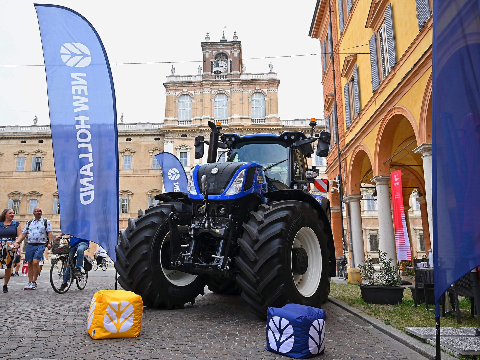A large blue New Holland tractor is displayed on a cobblestone street in front of historic buildings with arched windows and a clock tower. Blue New Holland flags and branded cube cushions are placed around the tractor. People walk and ride bicycles nearby, enjoying the outdoor event.