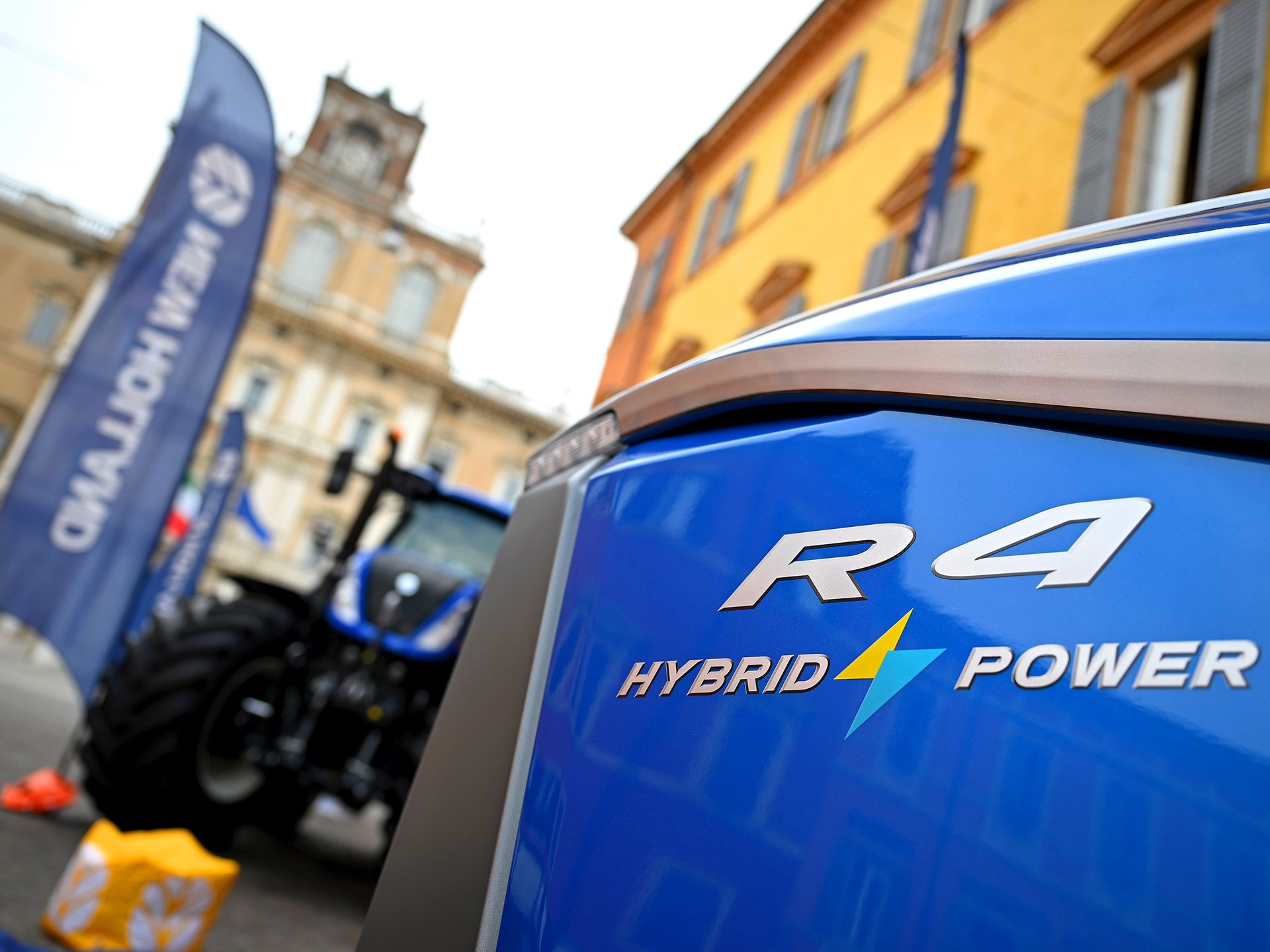 Close-up view of the front side of a blue R4 Hybrid Power tractor with a blurred background showing another tractor, flags, and historic buildings in an outdoor urban setting.