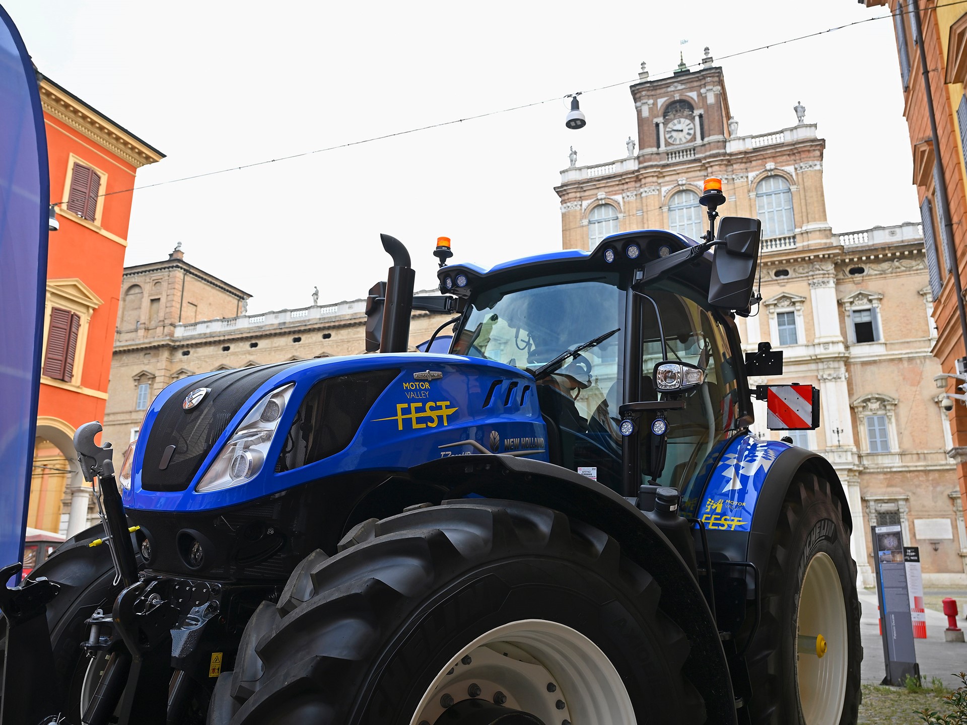 Blue New Holland tractor with "Motor Valley Fest" branding displayed in an urban setting with historic buildings in the background, under a clear sky.