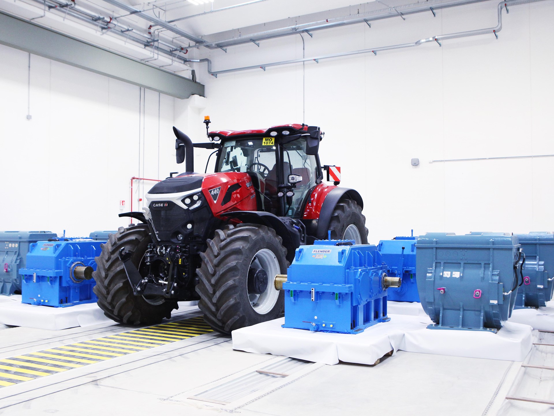 A red Case IH tractor is positioned indoors on a testing platform surrounded by several large blue industrial gearboxes and machinery components, all set in a clean, well-lit white room with visible ceiling pipes and safety markings on the floor.