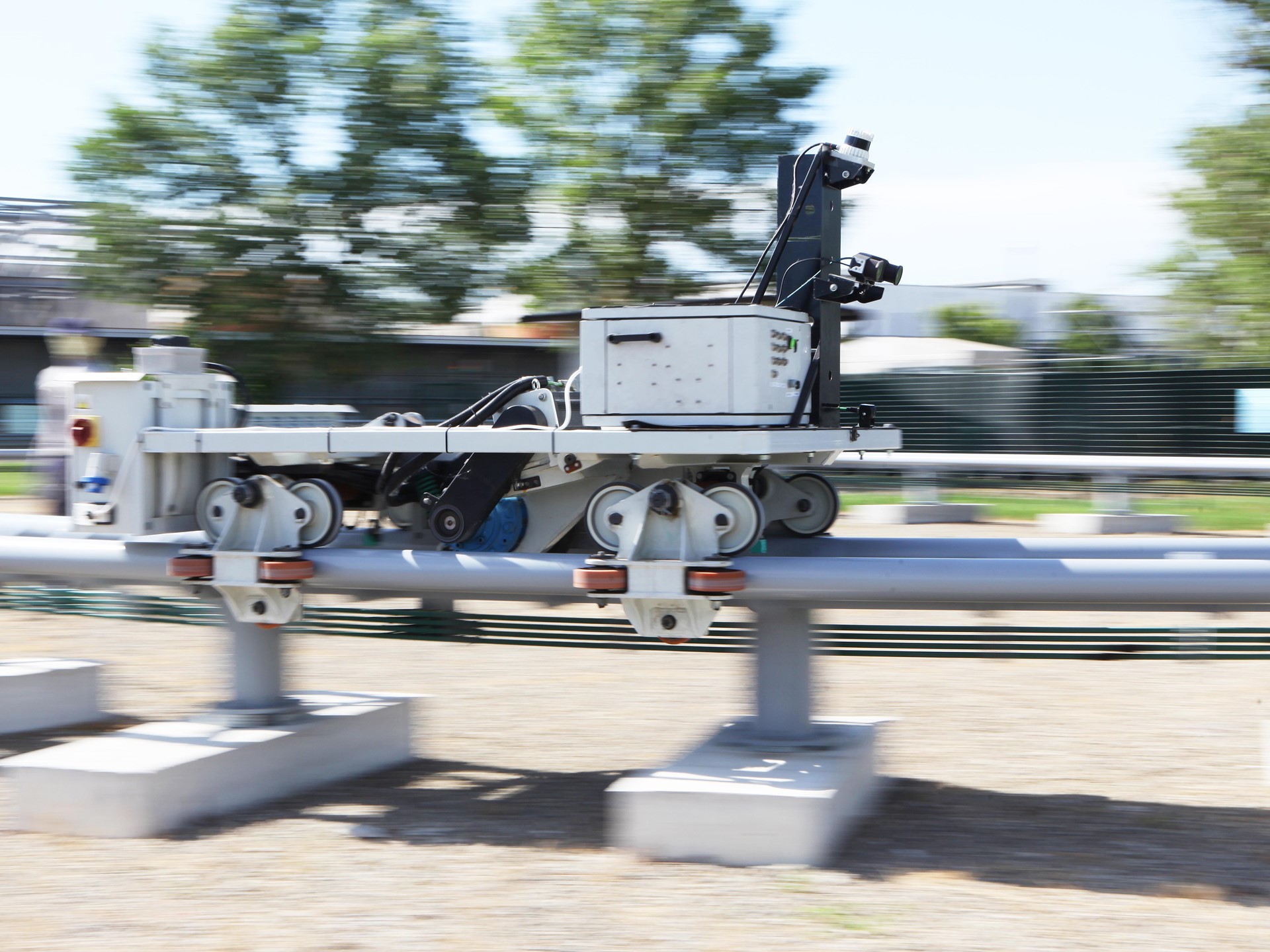 A mobile robot equipped with cameras and sensors is moving along a metal rail outdoors, with blurred trees and buildings in the background indicating motion.