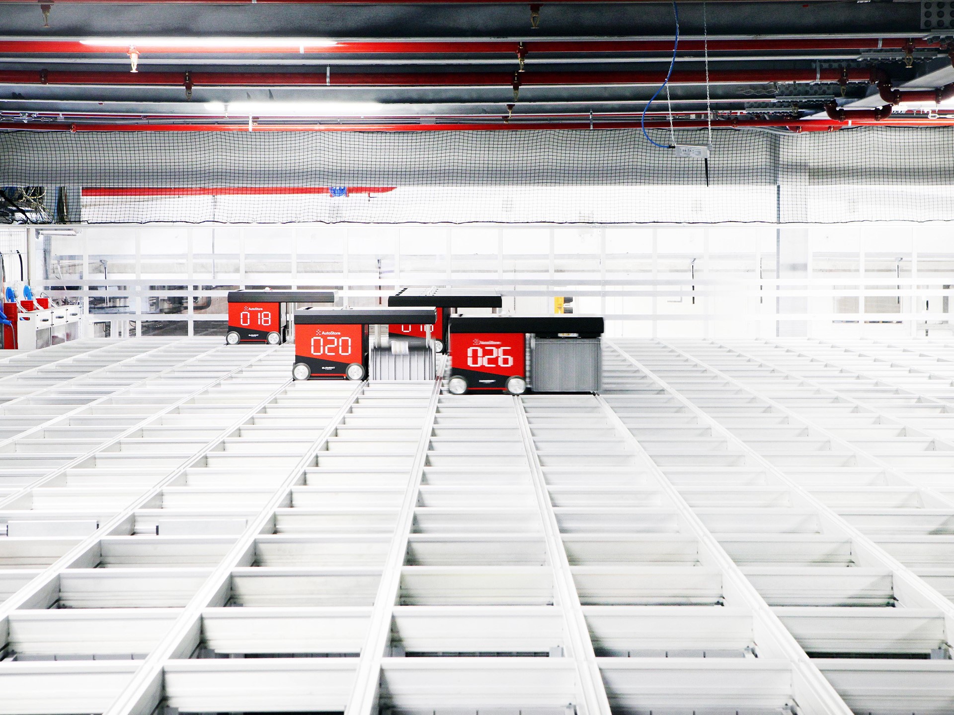 A high-tech automated warehouse with red robotic carts numbered 018, 020, and 026 moving along white grid-like storage shelves under a ceiling with red pipes and industrial lighting.