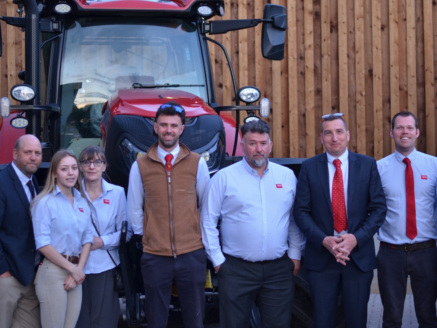 A group of eight professionally dressed people standing in front of a large red tractor and a wooden wall, posing for a group photo. Most individuals are wearing light blue shirts, some with red ties, and one person is wearing a brown vest.