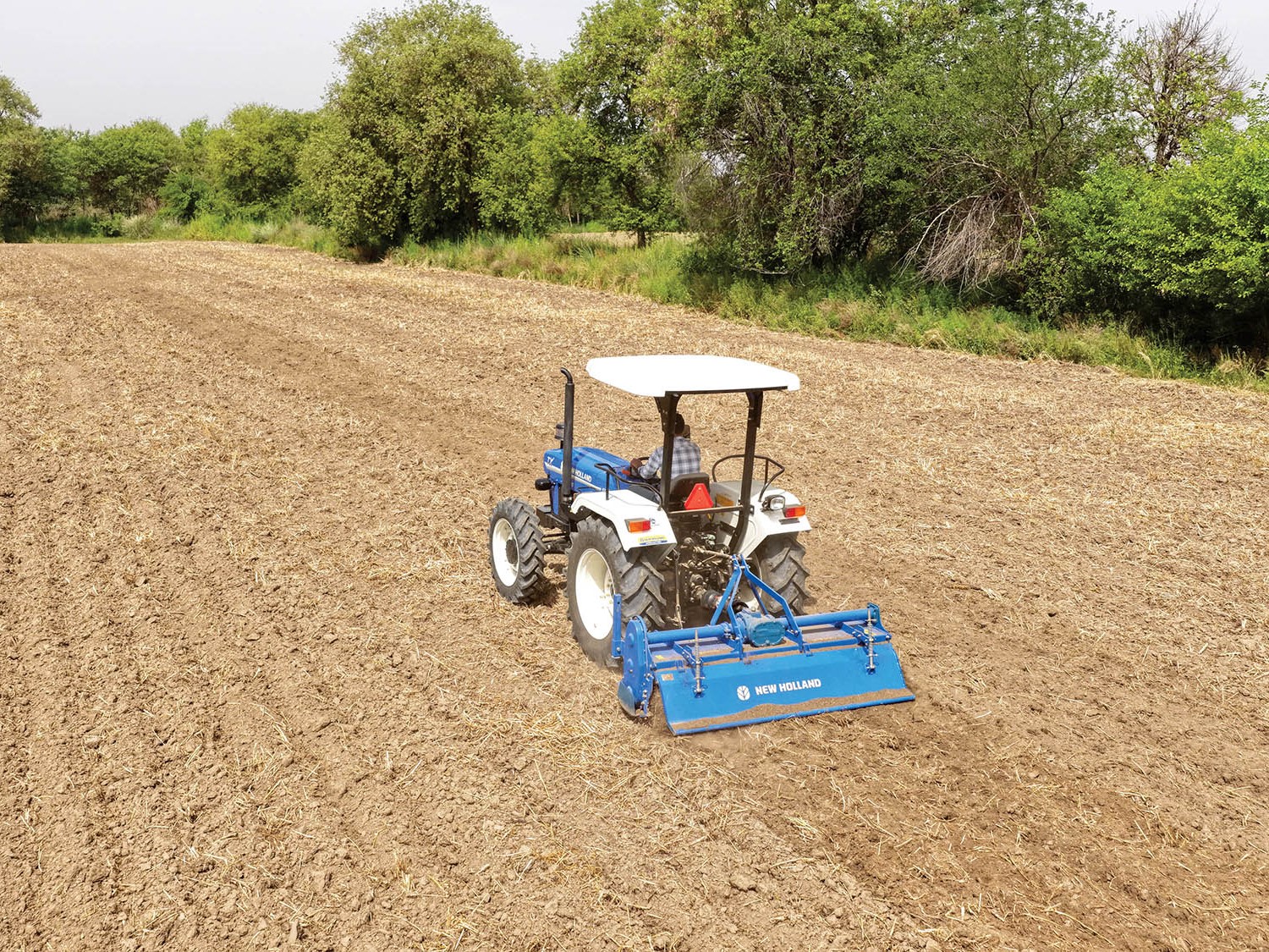 A blue and white New Holland tractor is tilling a large, dry, and freshly plowed field surrounded by green trees under a clear sky.