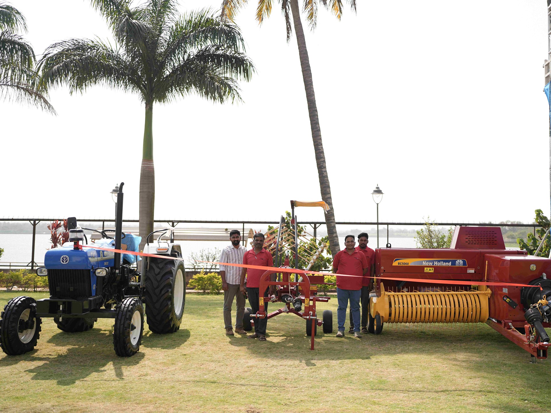 A group of six men standing outdoors on grass near agricultural machinery, including a blue tractor on the left and a large red New Holland hay baler on the right, with palm trees and a body of water in the background under a clear sky.