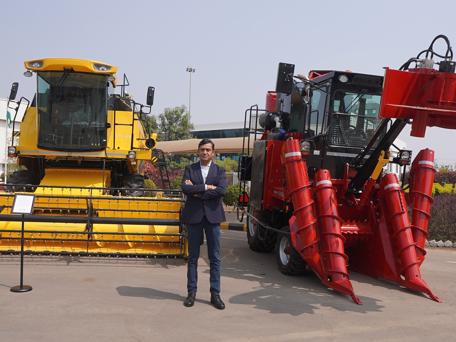 A man in a dark blazer and jeans stands with arms crossed between two large agricultural machines, one yellow and one red, parked on a paved area outside a building under a clear sky.