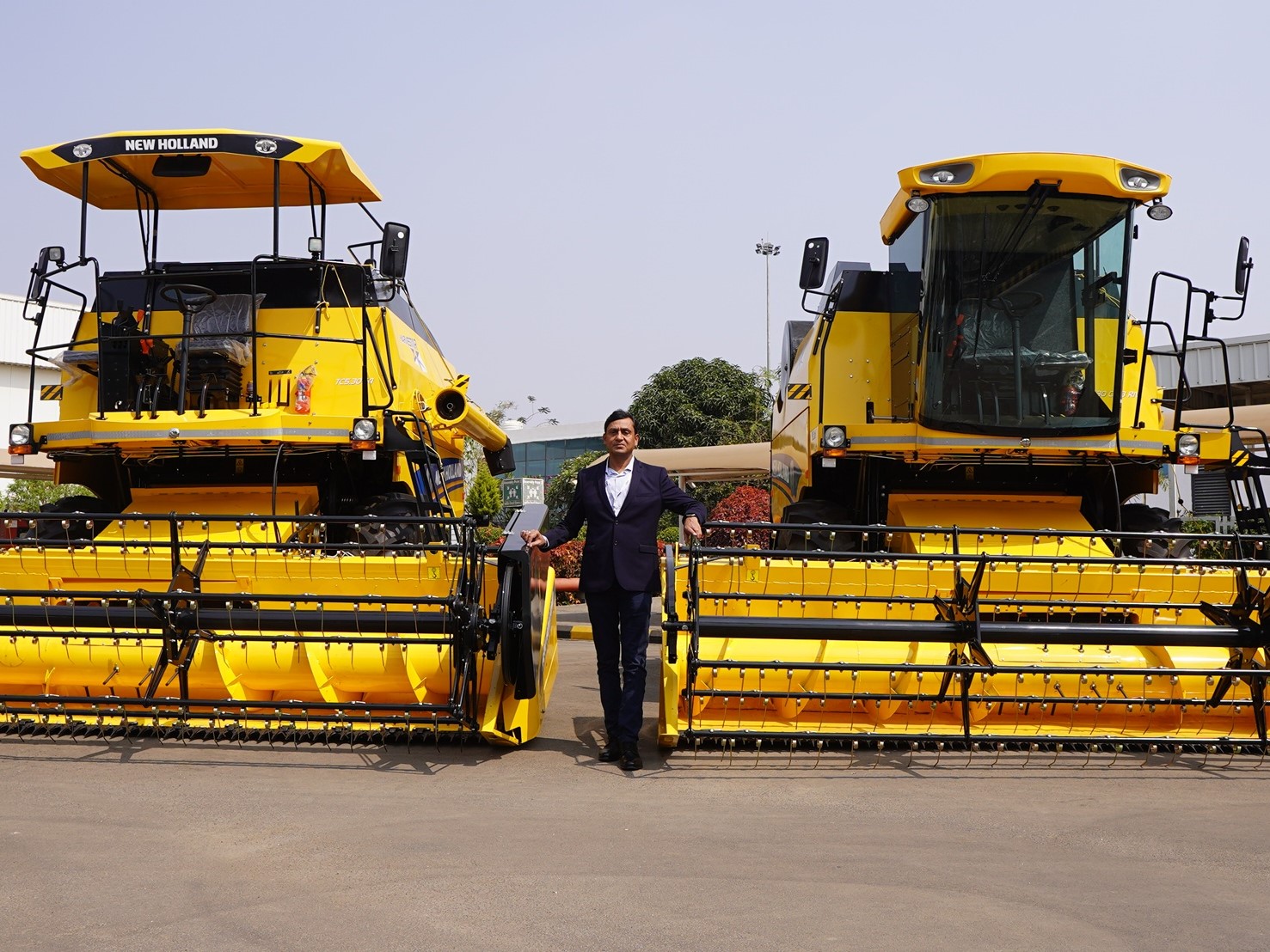 A man in a dark suit standing between two large yellow New Holland agricultural combine harvesters parked outdoors on a paved surface, with a building and greenery in the background under a clear sky.