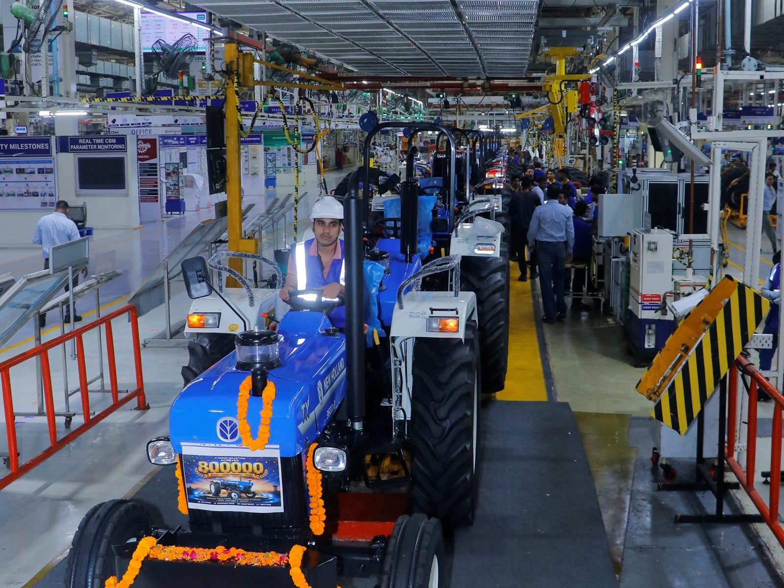 A blue tractor decorated with orange marigold garlands is being driven by a worker wearing a white helmet and reflective vest inside a large, well-lit industrial factory. The factory floor is busy with machinery, equipment, and other workers in the background, indicating a manufacturing or assembly line environment.