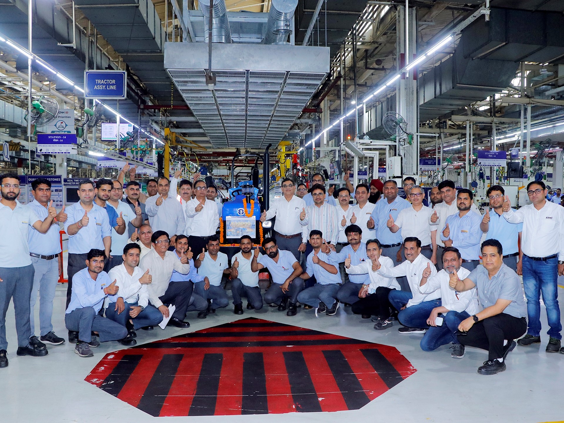 A large group of factory workers and engineers posing together inside a manufacturing facility, all giving thumbs-up gestures. They are standing and squatting around a blue tractor assembly line area, with industrial equipment and machinery visible in the background. The environment is well-lit with overhead lights and organized workstations.