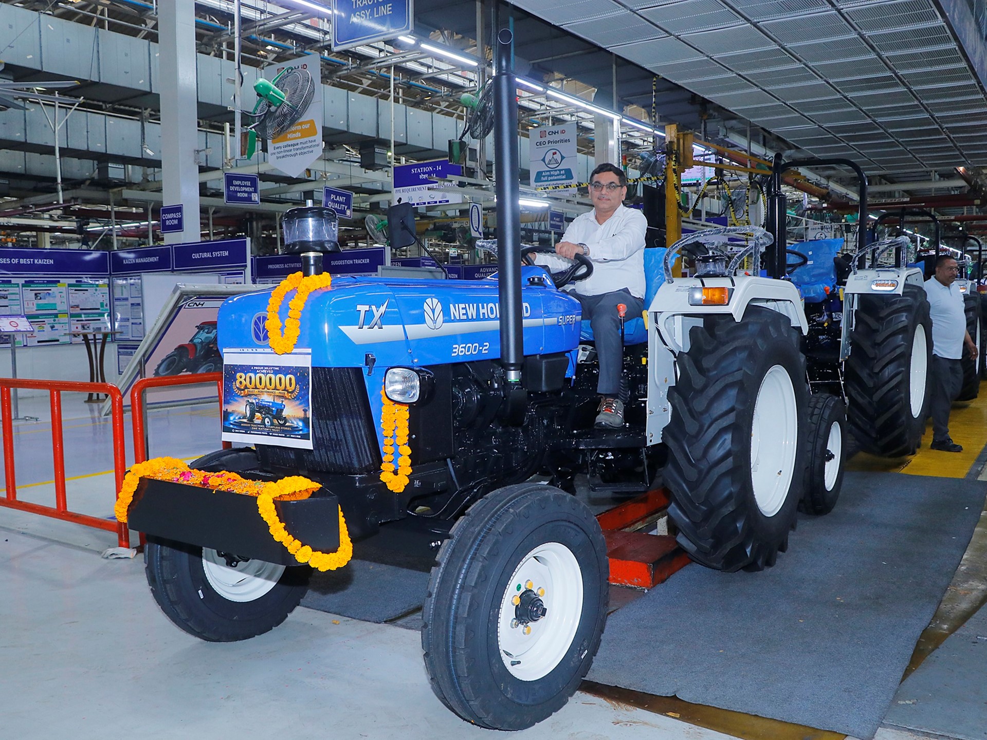 A man sitting on a blue New Holland TX 3600-2 tractor decorated with orange marigold garlands inside a factory assembly line. The tractor has a sign indicating "800000" units produced. Several similar tractors are lined up behind it, and factory workers are visible in the background. The setting is a well-lit industrial environment with various signs and equipment.