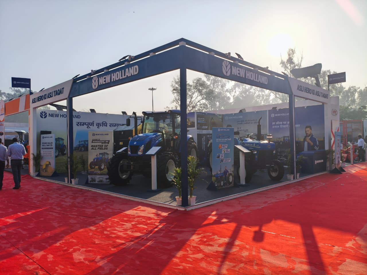 Outdoor exhibition booth of New Holland showcasing blue tractors on a red carpeted area with banners and informational displays, under a clear sky with sunlight and trees in the background.