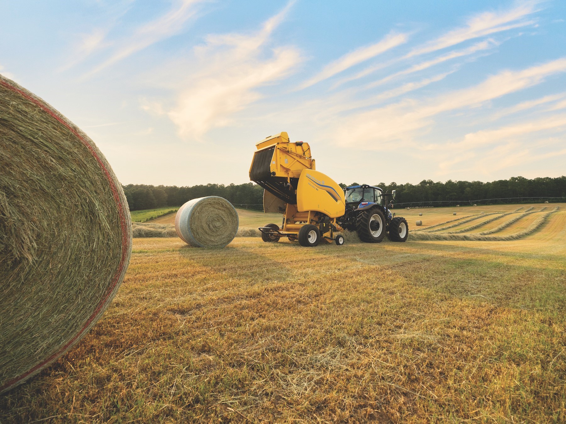 A blue tractor with a yellow hay baler attachment is working in a large open field during sunset, creating round hay bales. One large hay bale is in the foreground on the left, with more hay bales scattered across the field and a line of trees in the background under a partly cloudy sky.
