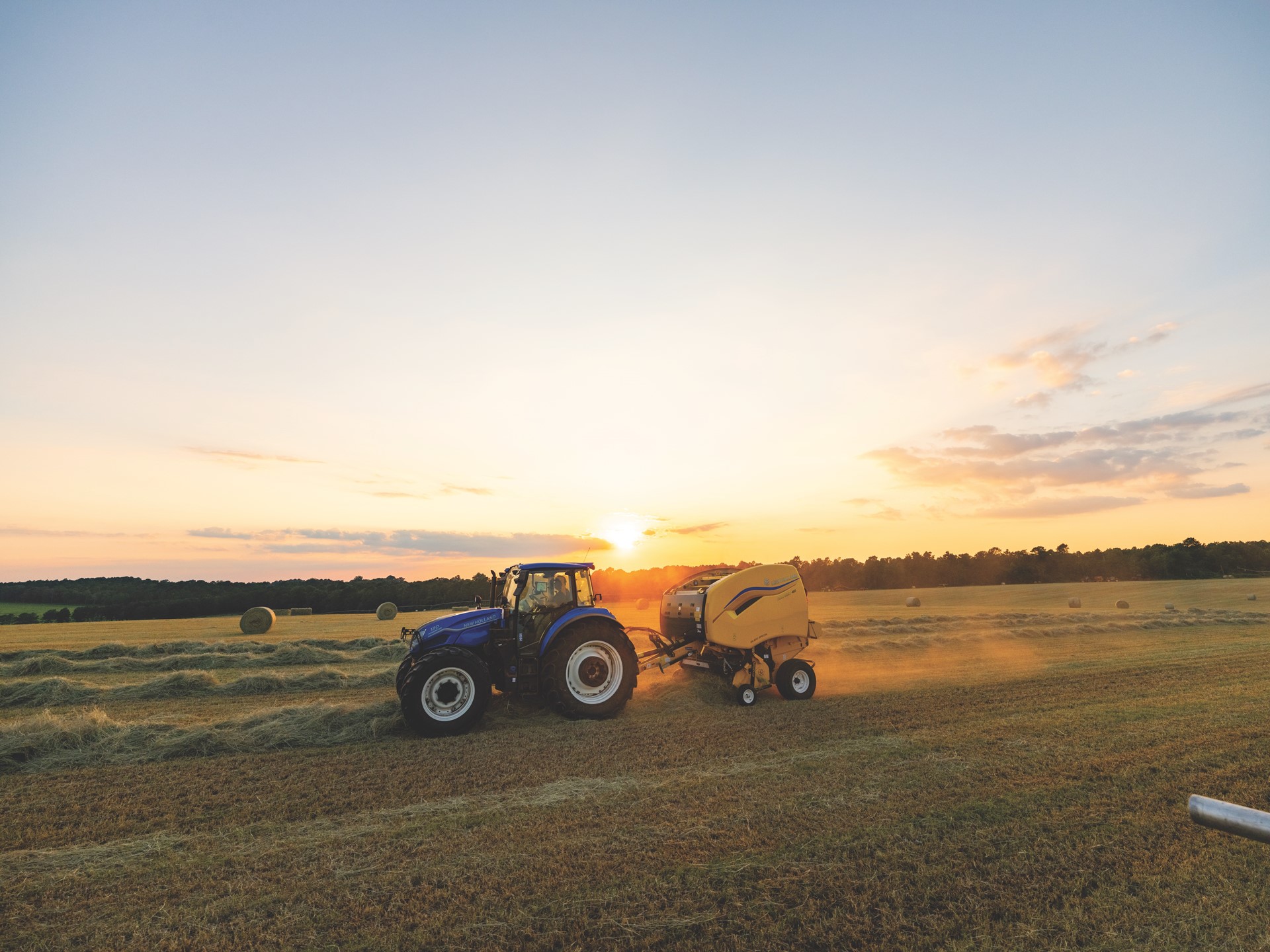 A blue tractor pulling a yellow baler machine in a large open field during sunset, with round hay bales scattered across the field and a clear sky with soft clouds in the background.