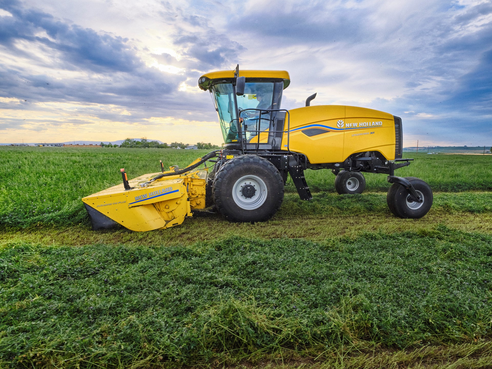 A yellow New Holland agricultural harvester machine is parked on a green field under a partly cloudy sky during daylight, with freshly cut crops visible on the ground.
