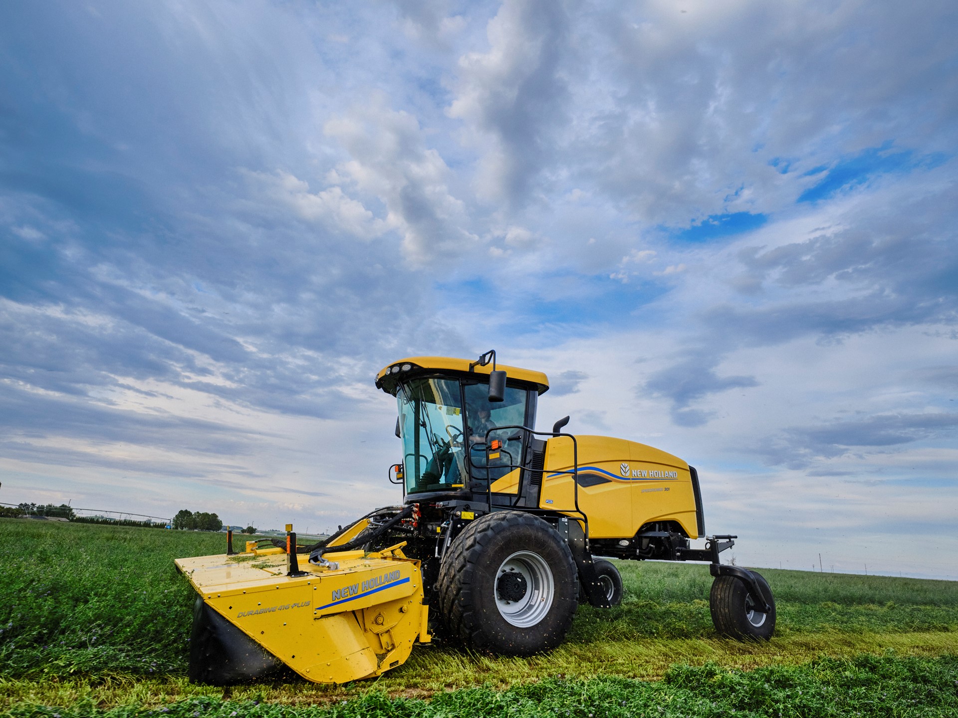 A yellow agricultural mower machine cutting green grass in a vast field under a partly cloudy blue sky during daytime.