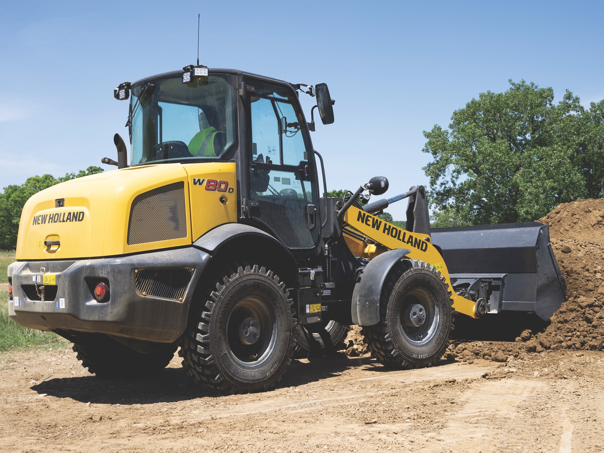 Yellow New Holland W80D wheel loader with a front bucket filled with dirt, parked on a construction site with trees and a clear blue sky in the background.