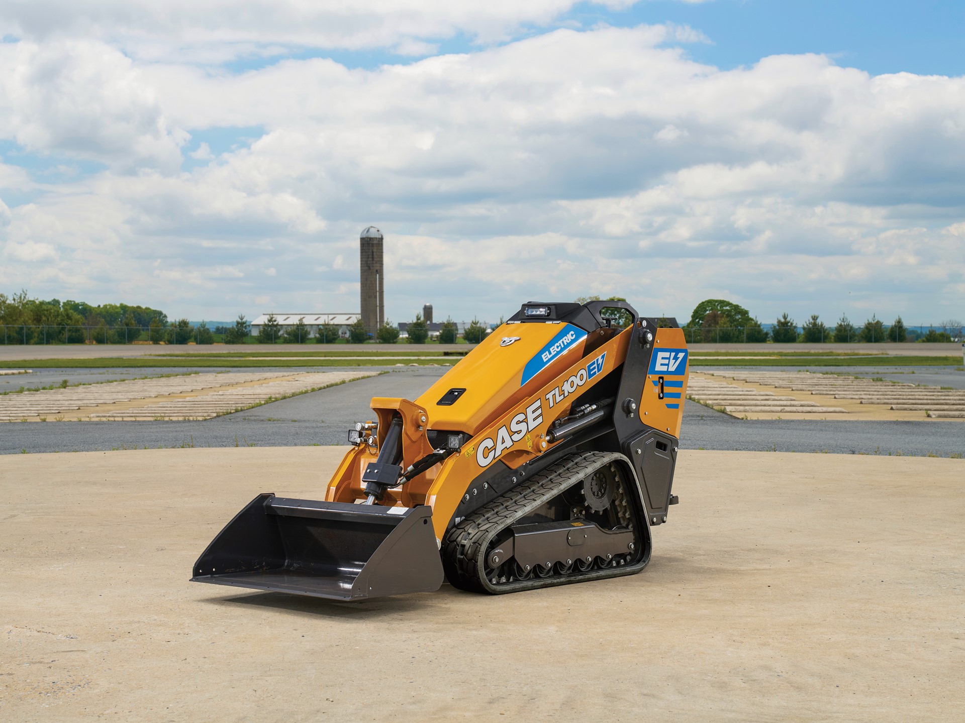 A yellow CASE TL100EV electric mini track loader with black tracks is parked on a concrete surface outdoors under a partly cloudy sky with a rural landscape and silo visible in the background