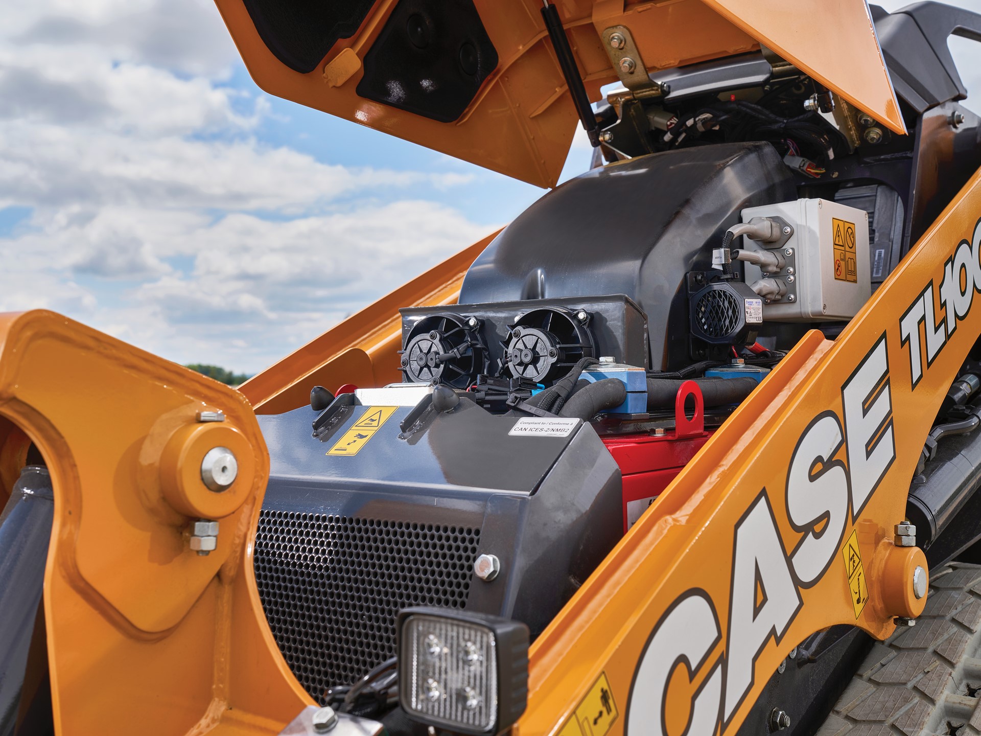 Close up view of the open engine compartment of a yellow CASE TL100EV electric mini track loader under a partly cloudy sky