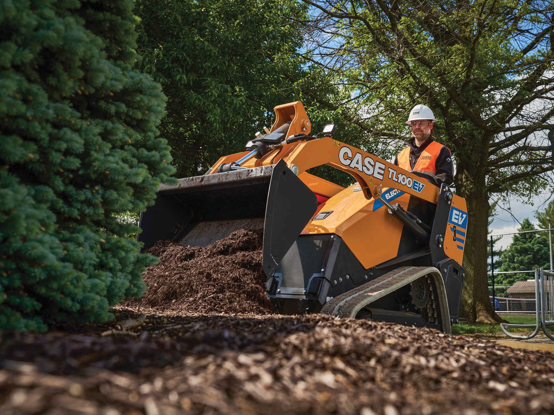 A worker wearing a white hard hat and orange safety vest operates a yellow CASE TL100EV electric mini track loader spreading mulch around a landscaped area with trees and greenery in the background