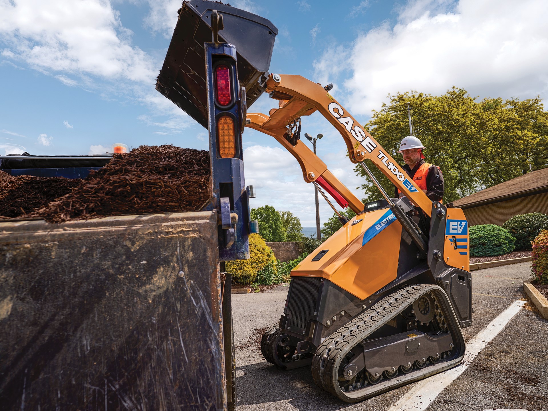 A worker wearing a white hard hat and orange safety vest operates a yellow CASE TL100EV electric mini track loader dumping mulch into a large container on a sunny day with blue sky and scattered clouds