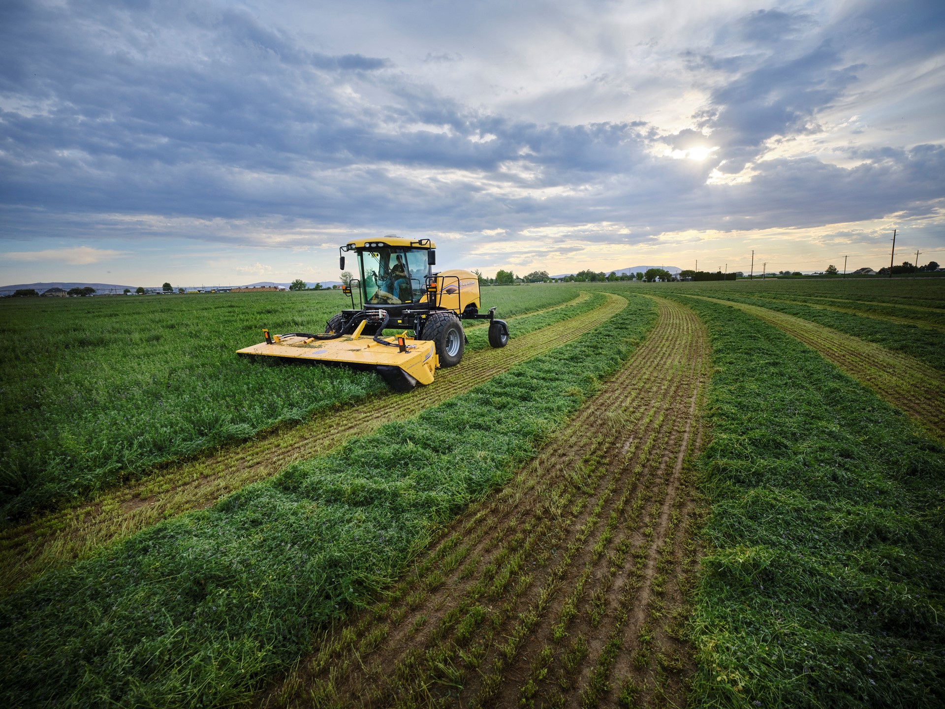 A yellow agricultural tractor mowing a large green field under a cloudy sky with the sun partially visible, creating neat rows of cut grass on the farmland.