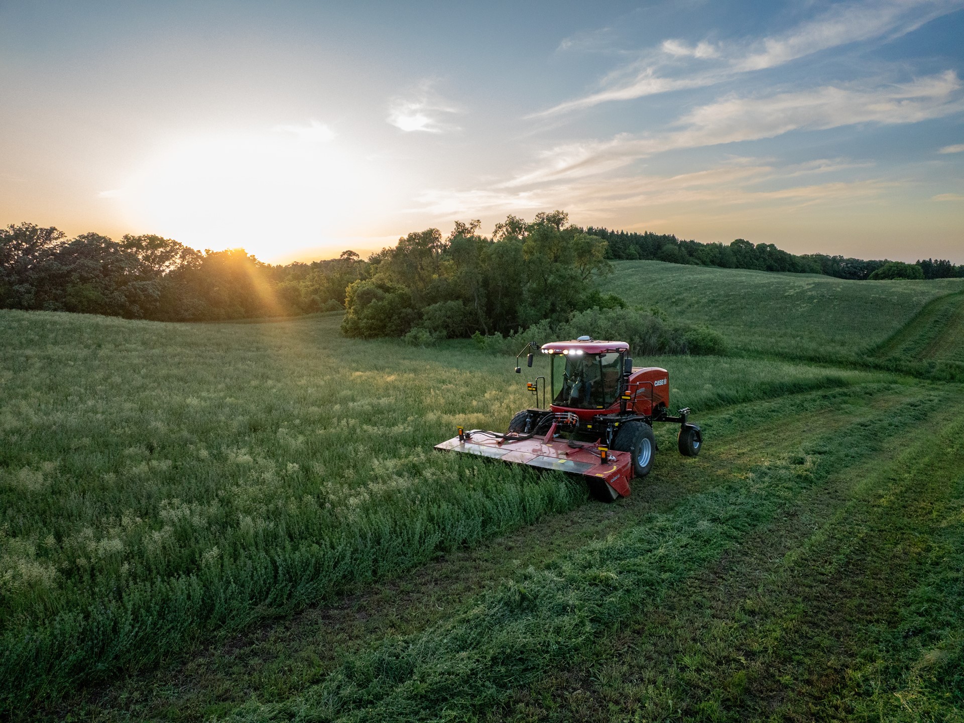 A red agricultural mower cutting grass in a large green field during sunset, with trees and rolling hills in the background under a partly cloudy sky.