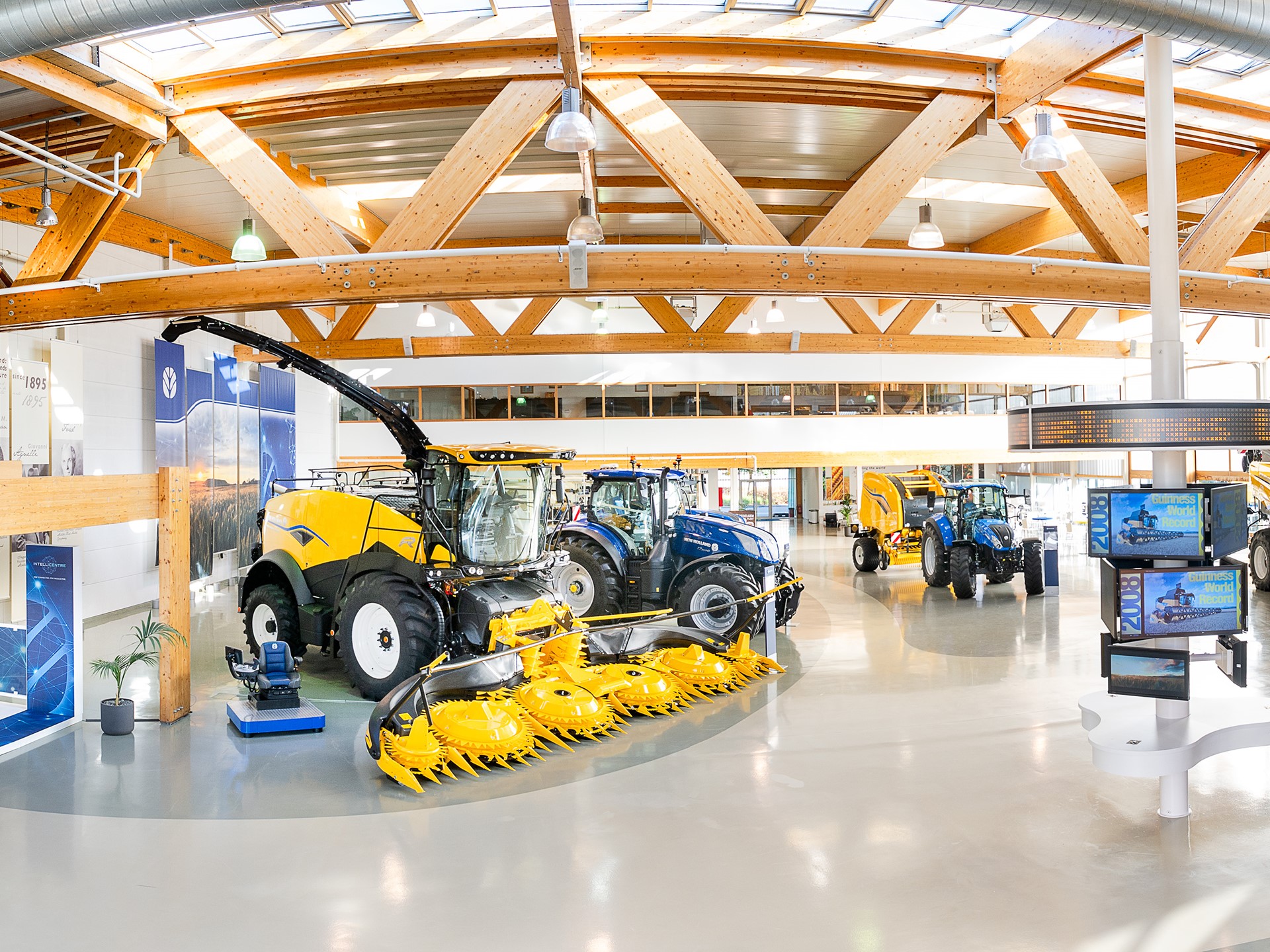 Interno di un moderno showroom agricolo con macchinari agricoli di grandi dimensioni, tra cui trattori blu, mietitrebbie gialle e attrezzature per la raccolta, illuminato da ampie finestre e travi in legno a vista.