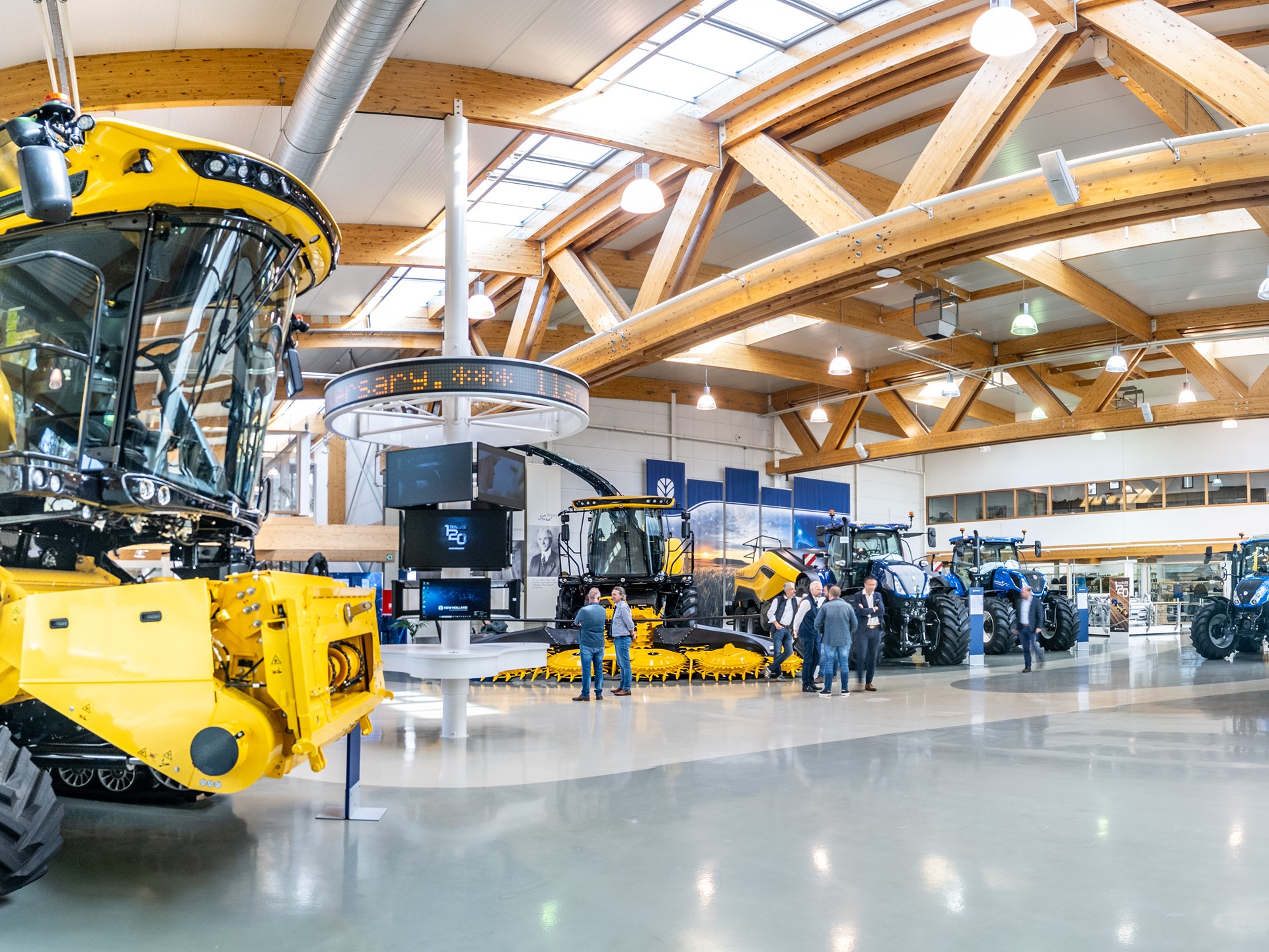 Interno di un grande showroom con macchinari agricoli moderni di colore giallo e blu, tra cui mietitrebbie e trattori, con alcune persone che osservano e discutono vicino ai veicoli, struttura con travi in legno e ampie finestre che lasciano entrare molta luce naturale.