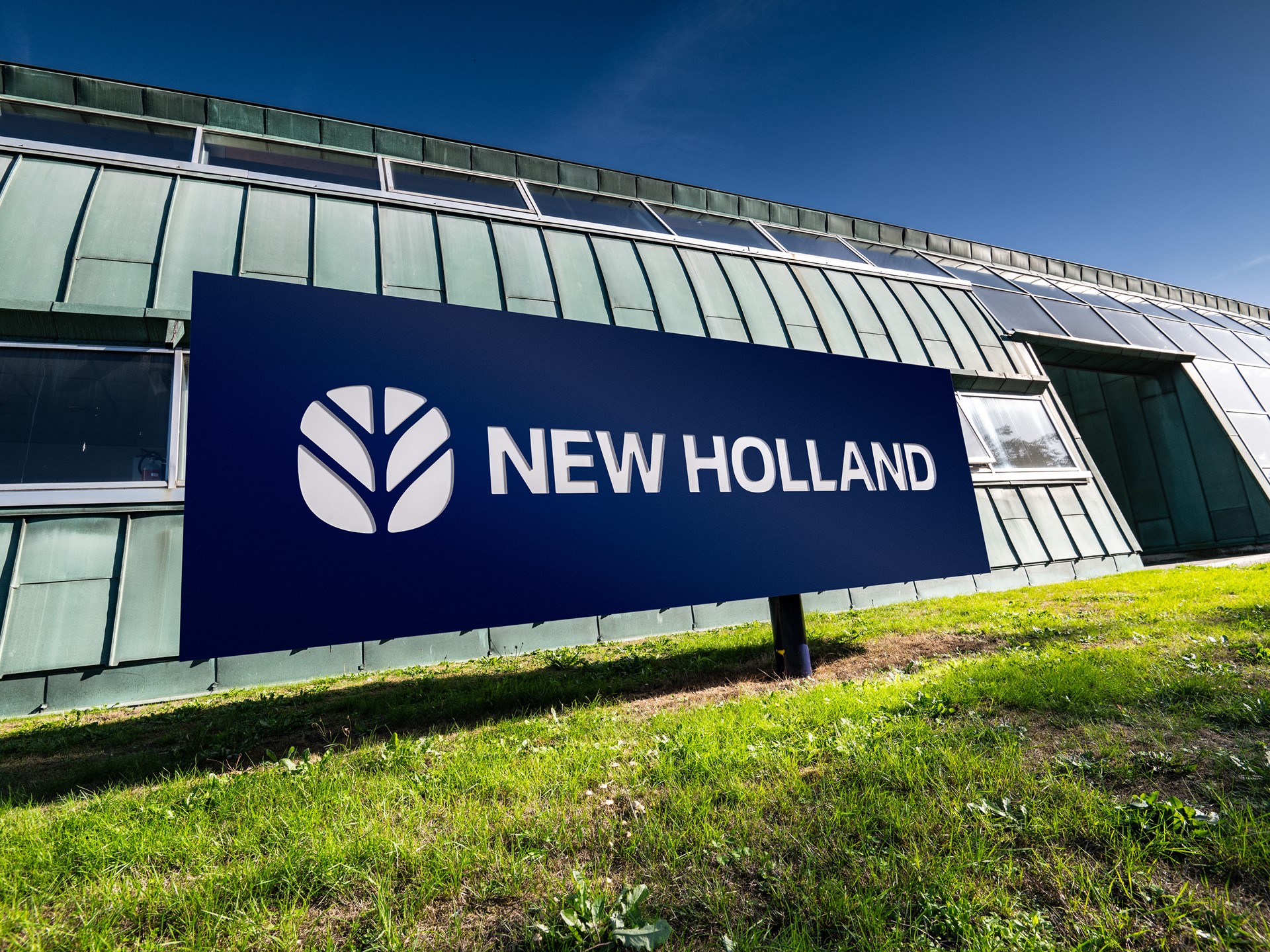 Insegna blu con il logo e la scritta "NEW HOLLAND" davanti a un edificio moderno con pareti in metallo verde e finestre, su un prato verde sotto un cielo sereno.
