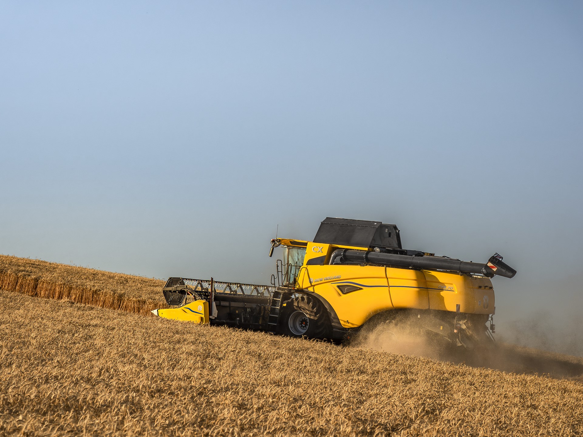 A yellow combine harvester working on a golden wheat field under a clear blue sky, harvesting crops and kicking up dust as it moves along a sloped terrain.
