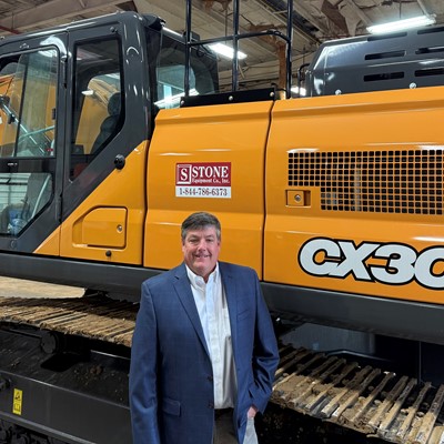 A man in a suit stands in front of a large orange Case CX300E excavator inside a warehouse. The excavator features a prominent logo and is positioned on a track, showcasing its size and design.