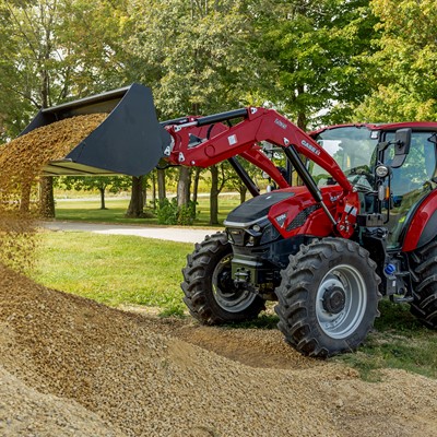 A red tractor with a front loader is dumping a pile of gravel onto the ground in a green outdoor setting. The tractor is positioned on a dirt path surrounded by trees, showcasing a sunny day. The loader is raised, and gravel is cascading down from the bucket.