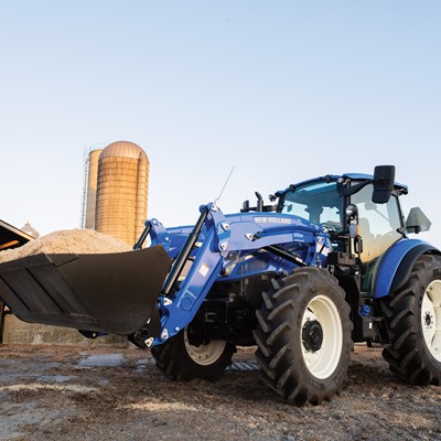 A blue New Holland tractor with a front loader is positioned on a farm, carrying a large bucket of material. In the background, a silos and a barn are visible under a clear sky, indicating a rural agricultural setting. The tractor's tires are large and rugged, suitable for farm work.