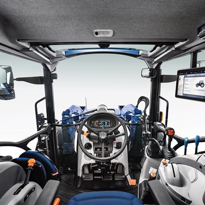 Interior view of a modern tractor cabin, featuring a steering wheel, control levers, and a digital display screen. The cabin is designed for operator comfort and efficiency, with ergonomic seating and various control buttons on the dashboard. Large side mirrors are visible, enhancing visibility for the driver.