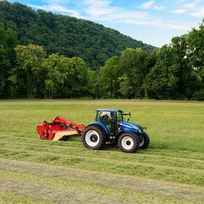 A blue tractor with a red mower attachment is cutting grass in a lush green field. In the background, rolling hills covered with trees are visible under a clear blue sky. The scene captures a peaceful rural landscape, emphasizing agricultural activity.