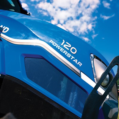 Close-up view of a blue New Holland tractor with the model name "120 Powerstar" prominently displayed. The image captures the tractor's sleek design against a backdrop of a clear blue sky with scattered clouds, highlighting its modern features and agricultural utility.