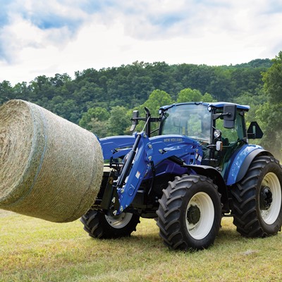 A blue New Holland tractor is lifting a large round hay bale in a grassy field. The background features lush green trees and a cloudy sky, indicating a rural farming setting. The tractor's front loader is engaged, showcasing its capability for handling heavy loads.